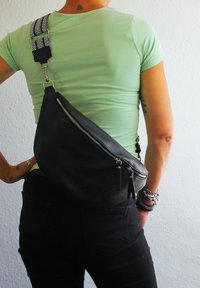 Zwarte leren schoudertas met een textuurafwerking, ritscompartiment en een gestreepte verstelbare schouderband. Draagtas op de rug, gecombineerd met donkere jeans.