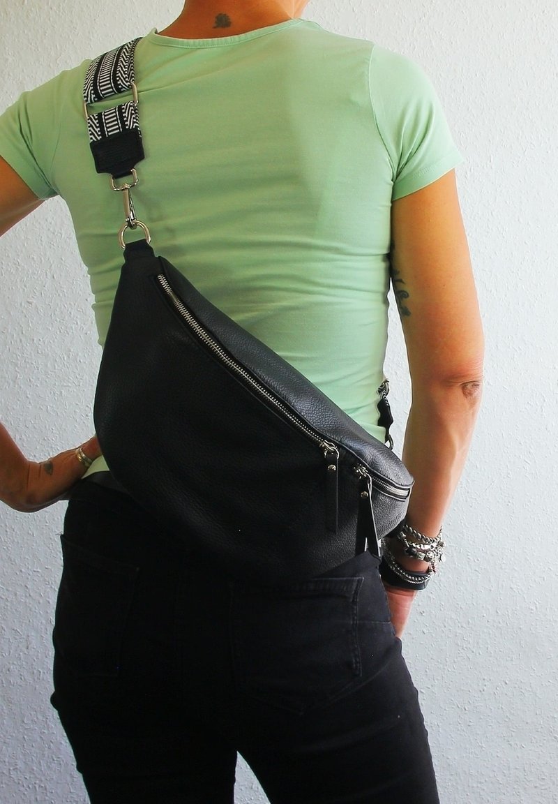 Zwarte leren schoudertas met een textuurafwerking, ritscompartiment en een gestreepte verstelbare schouderband. Draagtas op de rug, gecombineerd met donkere jeans.