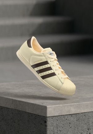 Kremowy sneaker Adidas Superstar z ciemnobrązowymi paskami, unoszący się nad szarą betonową krawędzią w miejskim otoczeniu.