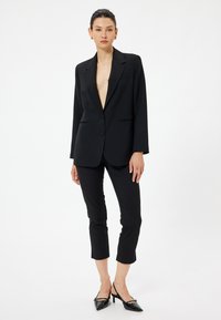 Svart skräddarsydd blazer med djup V-ringning, enkel knappstängning och framfickor, som kombineras med figurnära svarta capribyxor och glänsande svarta klackar.