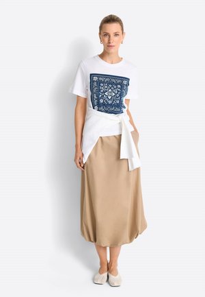 Donna in piedi che indossa una t-shirt bianca con stampa quadrata floreale blu, maglione bianco annodato in vita, gonna midi beige e scarpe crema senza lacci.
