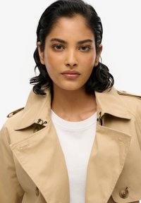 Beżowy trencz z wyraźnym kołnierzem i guzikami, noszony na białej koszulce; gładka tekstura i klasyczny design.