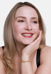 Bracciale in oro su un polso, pelle liscia, capelli castano chiaro, pelle luminosa e espressione vivace con trucco leggero.