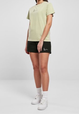 Hellgrünes Baumwoll-T-Shirt mit Rundhalsausschnitt und kurzen Ärmeln, kombiniert mit schwarzen Shorts. Weiße Schuhe und Socken vervollständigen das Outfit.