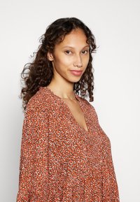 Vestido de manga longa com decote em V, em laranja vibrante, com um padrão abstrato em branco e preto. Tecido leve com um corte solto e decote com babados.