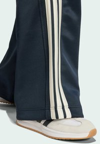 Pantalones deportivos navy con tres franjas verticales claras a los lados, combinados con zapatillas deportivas ligeras que tienen acentos en negro y blanco.