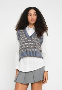 Joven con cabello rizado que lleva un chaleco de punto a rayas azul y gris sobre una camisa blanca con cuello y una falda corta plisada gris.