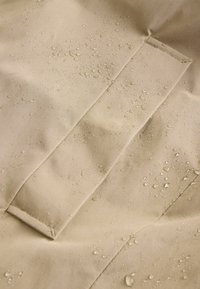 Beige vedenkestävä kangas, jossa on nähtävissä vesipisaroita. Teksturoitu pinta sisältää saumoja ja läppäyksikön. Yhteenvetona pehmeä ja sileä ilme.