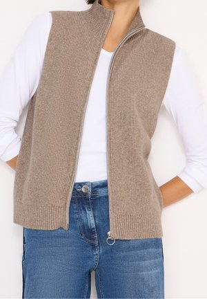 Vrouw draagt een beige gebreid vest met ritssluiting over een wit shirt met lange mouwen en een blauwe spijkerbroek, met haar handen achter haar rug.