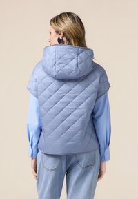 Lichtblauw gewatteerd vest met capuchon, voorzien van ruitjesstiksels, korte mouwen, gecombineerd met een lichtblauw shirt met lange mouwen en denim jeans.