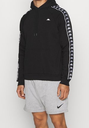 Mężczyzna w czarnej bluzie z kapturem Kappa z taśmą z logo na rękawach oraz szarych szortach Nike z czarnym logo Swoosh.