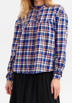 Femme portant une blouse à carreaux rose, bleue et blanche à manches longues et une jupe noire.