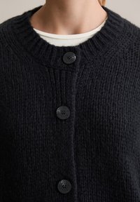 Cardigan a maglia blu scuro con collo a costine, caratterizzato da quattro bottoni neri e un tessuto strutturato. Vista ravvicinata che mette a fuoco il colletto.