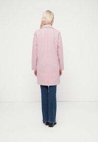 Ροζ μάλλινο παλτό με γιακά, μακριά μανίκια, λεπτομέρεια στην πλάτη με σκίσιμο και ευθεία γραμμή. Συνδυάζεται με λευκά τζιν καμπάνα και μαύρες μπότες με τακούνι.