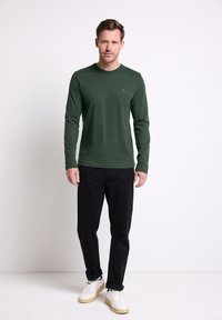 Zielona koszula z długim rękawem wykonana z miękkiej, fakturowanej tkaniny, zestawiona z czarnymi spodniami i białymi trampkami. Prosty okrągły dekolt, mały detal z logo.