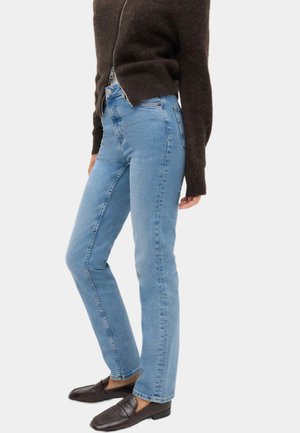 Person iført lyseblå straight-leg jeans, en mørkebrun lodden lynlåstrøje og mørkebrune loafers, der står i siden.