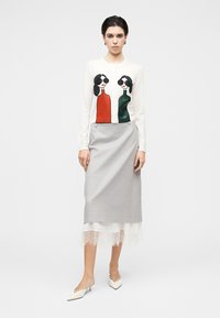 Cardigan blanc avec un motif graphique de deux femmes en rouge et vert, associé à une jupe crayon grise avec une bordure en dentelle à l'ourlet. Chaussures blanches à bouts pointus.