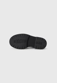 Suela de goma negra con surcos profundos para tracción. Nombres de marca en relieve "STEVE MADDEN" y "WADDEN" presentados de manera prominente. Textura suave.
