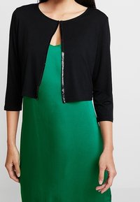 Musta lyhythihainen neuletakki, jossa on kolme neljännesosaa hihat ja helmiäispainamalla koristeltu etuläppä. Päällä vihreä mekko. Sileä kankaan rakenne.