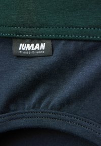 Svart klädlapp med vit text "IUMAN intimissimi uomo" sydd mellan grön och marinblå tyg.