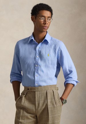 Férfi kerek szemüveggel, világoskék gombos inggel, feltűrt ujjakkal és magas derekú kockás nadrágban, kezei a zsebekben.