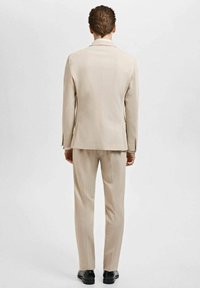 Beige kostym med en skräddarsydd kavaj och slim-fit byxor, tillverkad av ett mjukt tyg. Bakifrån syns en enkel slits och minimal detaljering.