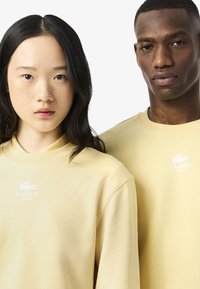 Sudaderas amarillas con el logo de Lacoste en blanco, con una textura suave y un corte relajado, llevadas por dos personas.