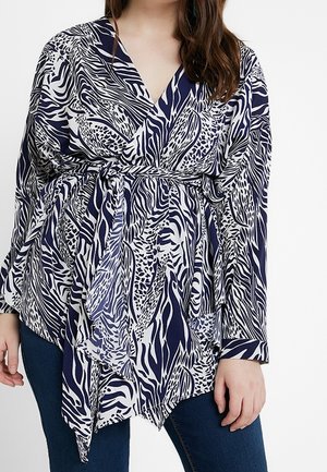 Kobieta ubrana w granatowo-białą bluzkę kopertową w zwierzęcy wzór z długimi rękawami i wiązanym paskiem, połączoną z ciemnoniebieskimi dżinsami.