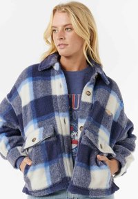 Blonde vrouw in een blauw en wit geruite fleecejack over een marineblauwe sweatshirt, met handen in de zakken van het jack, kijkt naar rechts.