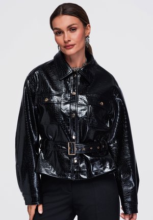 Femme portant une veste ceinturée noire brillante à motif croco, avec un devant boutonné et des boucles d’oreilles décoratives sur un fond uni.