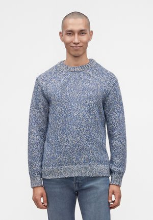 Mand med kort hår iført en blå- og hvid strikket sweater og blå jeans, stående mod en ensartet lys baggrund.