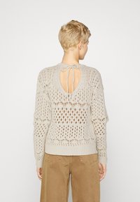 Pull en tricot beige clair avec un design clé au dos et une fermeture à lien, motif en openwork texturé, et des poignets et un ourlet côtelés.
