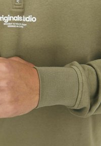 Olivgrön långärmad skjorta med ribbade ärmslut och dragkedja vid kragen. Har en vit textlogga på bröstet. Mjuk stickad tygtextur.