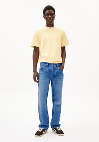 Gelbes Baumwoll-T-Shirt mit Rundhalsausschnitt, kombiniert mit blauen Jeans. Die Jeans haben einen geraden Schnitt und sichtbare Nähte. Es werden schwarze Sneakers getragen.