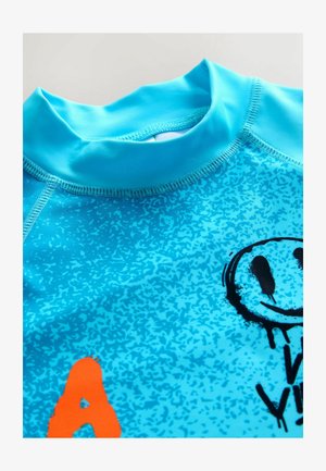 Jasnomodrý rashguard s hladkou textúrou, krátkym vysokým golierom a grafickými vzormi v čiernej a oranžovej farbe.