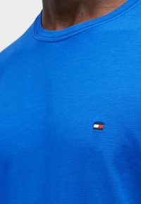 Κοντινό πλάνο ενός ατόμου που φοράει φωτεινό μπλε μπλουζάκι με στρογγυλή λαιμόκοψη και μικρό ορθογώνιο κόκκινο, λευκό και μπλε λογότυπο στο στήθος.