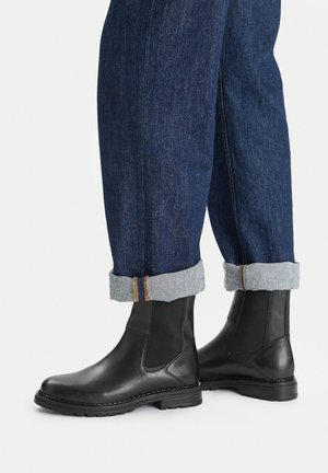 Czarne skórzane botki z elastycznymi panelami po bokach, gumowymi podeszwami i przeszyciami, w połączeniu z ciemnymi dżinsami z podwiniętymi mankietami.
