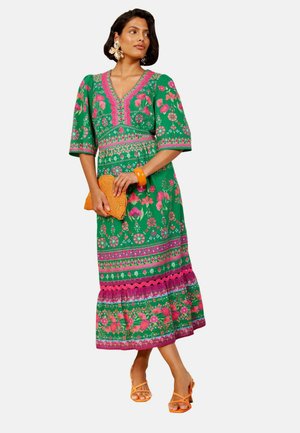 Kvinde iført en grøn blomsterprintet midi-kjole med pink accenter, der holder en orange vævet clutch, og som har orange sandaler og store øreringe på.