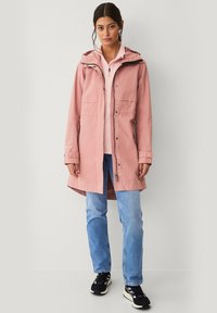 Chaqueta impermeable rosa con capucha, cierre de broche y cremallera, dos bolsillos delanteros y puños ajustables, combinada con vaqueros azul claros y zapatos negros.