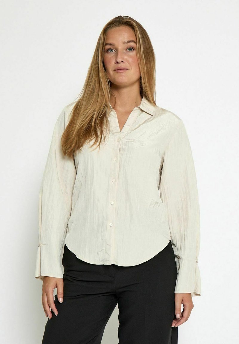 Vrouw met lang blond haar, gekleed in een crèmekleurige blouse met knopen en zwarte broek, staand tegen een effen witte achtergrond.