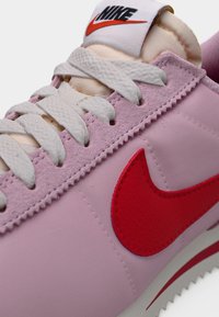 Nike-sneaker med en ljusrosa slät ovandel, vita skosnören, röd Swoosh-logga och vadderad vit tunga. Har texturerade accenter och söm-detaljer.
