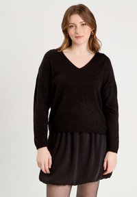 Black, knitted sweater with a V-neck and dropped shoulders, featuring a relaxed fit and ribbed hem, paired with a black dress.