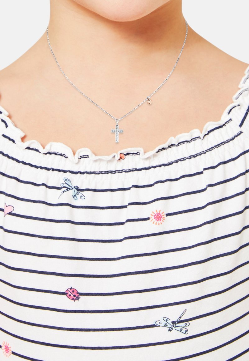 Collana in argento con pendente a croce decorato con strass e un piccolo charm a forma di cuore. Indossata sopra una maglietta bianca a righe con dettagli ricamati.