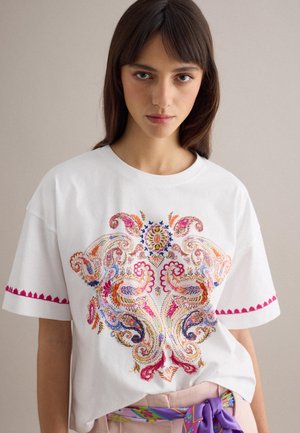 Jonge vrouw draagt een wit T-shirt met kleurrijke paisley-borduurwerk en roze bies aan de mouwen, gecombineerd met beige broek en een paarse riem met patroon.