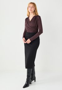 Haut à manches longues et col en V profond de couleur marron foncé avec tissu drapé, associé à une jupe crayon noire taille haute et des bottes noires à talons.