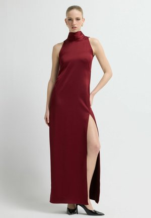 Bordeauxrode satijnen maxi-jurk met een hoge hals, een zijsplit, een nauwsluitende pasvorm en een gladde textuur. Model gecombineerd met zwarte hakken.