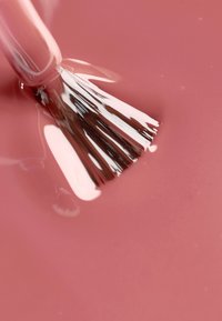 Nagellak met een romige, rozenroze finish. Een kwast wordt in de lak gedoopt, waarbij zachte haren en een reflecterende vloeibare textuur zichtbaar zijn.