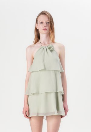 Hellgrünes, schichtweise gestaltetes Kleid mit Neckholder, das auf der linken Seite eine Stoffblüte hat. Hergestellt aus weichem, fließendem Material.