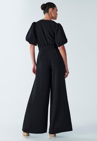 Zwarte jumpsuit met wijde pijpen en pofmouwen, getailleerde taille en een gladde textuur. Gouden hakken met enkelriempje maken de outfit compleet.
