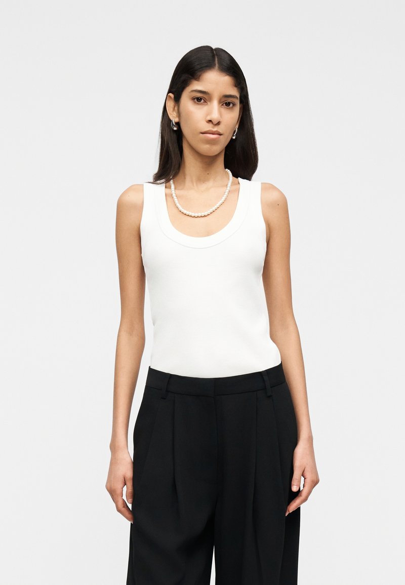 Joven mujer vestida con una blusa sin mangas blanca, pantalones negros de pierna ancha, pendientes de aro y un collar de perlas, posando contra un fondo liso.